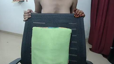 Media: Video of a topless person holding a green pillow on a gray chair, with a maroon curtain in the background.