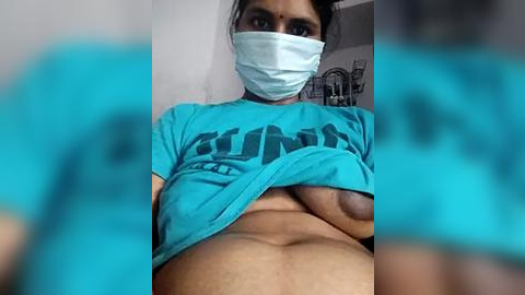 Media: A video of a pregnant woman with a blue mask, lifting her blue t-shirt to expose her bare chest and belly, taken indoors with blurred background.