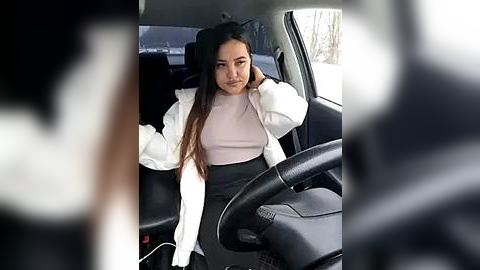 Media: Video of a young woman with long brown hair, wearing a white jacket over a pink top and black skirt, seated in a car, driving with her right hand.