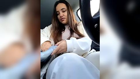 Media: Video of a young woman with long, straight brown hair, wearing a white hijab and light-colored top, sitting in a car, holding the steering wheel with a neutral expression.