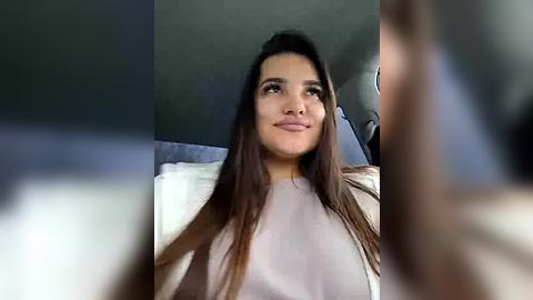 Media: Video of a young Latina woman with long, straight brown hair, wearing a light-colored sweater, smiling, and looking upward. Background features a dark wall and indistinct figures.