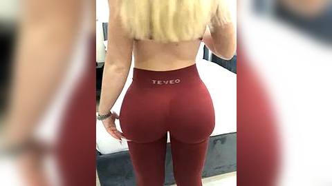 Media: Video of a blonde woman wearing maroon \"Tevco\" leggings, showing her toned buttocks and back, taken indoors with blurred background.