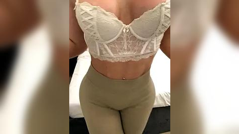 Media: Video of a woman in a beige lace bra and high-waisted leggings, standing in a bedroom with white sheets. The image is slightly blurred and cropped, focusing on her midsection.