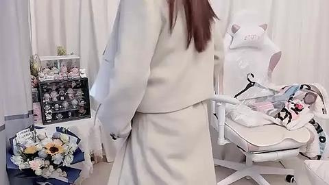 Media: Video of a woman in a white robe, standing in a plush, white room with a teddy bear on a chair, a floral arrangement, and a display case of plush toys.