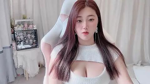 Media: Video of a fair-skinned woman with long, straight auburn hair, wearing a white cropped top that reveals her ample cleavage. She sits in a white gaming chair, surrounded by white curtains and a white bed.