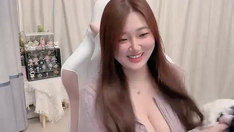 Media: Video of a smiling East Asian woman with long, straight brown hair, wearing a low-cut, purple top, sitting on a white gaming chair, in a softly lit, neutral-toned room.