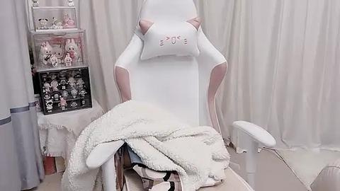 Media: A video of a white gaming chair with pink accents, draped with a soft, fluffy white blanket, surrounded by plush cat toys and a shelf with more toys.