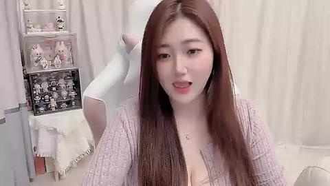 Media: Video of a young East Asian woman with long brown hair, wearing a lavender lace cardigan, smiling while adjusting her earbud. Background features a white curtain and a shelf with plush toys.