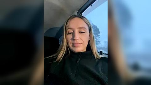 Media: Video of a blonde woman with light skin, wearing a black coat, smiling inside a car with snow outside the window.