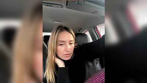 Media: Video of a young Caucasian woman with blonde hair, wearing a black sweater, seated in a car, looking tired with one hand on her head.