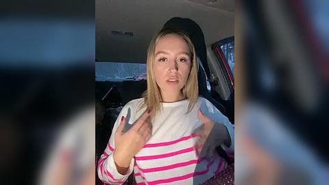 Media: Video of a young blonde woman with straight hair, wearing a white and pink striped sweater, gesturing with her hands in a car's backseat, blurred background.