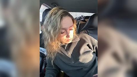 Media: Video of a blonde woman with shoulder-length hair, wearing a gray sweater, seated in a car, looking down. The car interior is dimly lit, and her expression is neutral.