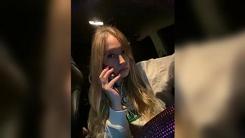 Media: Video of a young woman with long blonde hair, wearing a white sweatshirt, talking on her phone, seated in a dimly lit car.