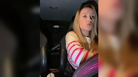 Media: Video of a young woman with long blonde hair in a car, wearing a striped sweater and looking serious. The background shows blurry, dark interior.