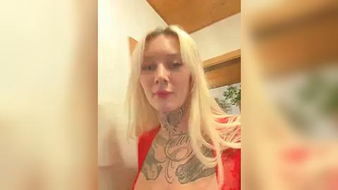 Media: Video of a blonde woman with long hair, wearing a red sweater, showcasing extensive neck tattoos. She stands indoors near a window with a wooden frame.