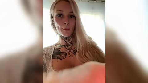 Media: Video of a tattooed, blonde woman with piercings, wearing a lacy top, looking into the camera. Background is blurry with a hint of red light.