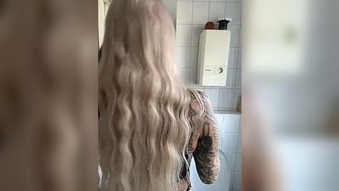 Media: Video of a person with long, wavy, platinum blonde hair, wearing a black top, standing in a tiled bathroom with a white heater and red objects on a shelf in the background.