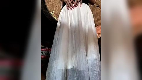 Media: Video of a woman wearing a white, flowing, pleated wedding gown, with hands placed on her waist, set against a rustic, woven background and blurred objects in the foreground.