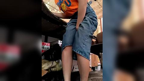 Media: Video of a person wearing blue denim overalls, squatting in a cluttered room with a table, a tent, and various objects in the background.