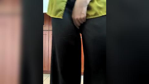 Media: A video of a person's lower body, wearing black pants and a mustard-yellow vest, standing in a kitchen with wooden cabinets and beige tiles. The person's hand is partially visible, adjusting their pants.