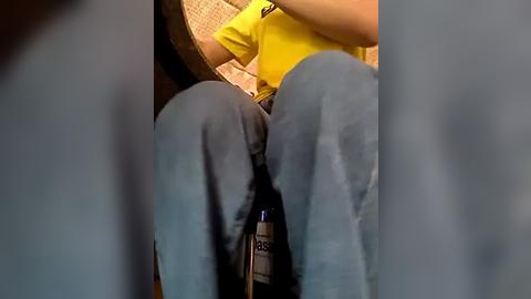 Media: A video showing a person's legs in blue jeans, a yellow shirt, and a bottle of beer on a wooden table, blurred background.