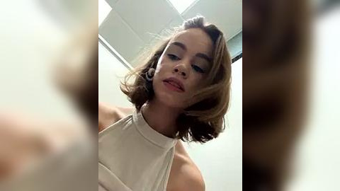 Media: Video of a young woman with light brown, shoulder-length hair, wearing a white halter-neck top, standing in a brightly lit room with white walls and ceiling tiles.