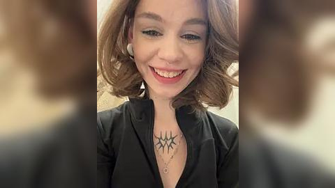 Media: Video of a smiling woman with shoulder-length, wavy, light brown hair, wearing a black blazer and white earbuds, featuring a tattoo of a black, geometric pattern on her chest.