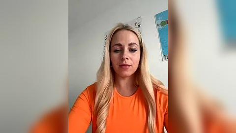 Media: A video of a fair-skinned, blonde woman with long hair wearing an orange top, standing in a white-walled room with a map and a bookshelf in the background.