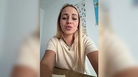 Media: A video of a young, fair-skinned woman with straight blonde hair, wearing a beige t-shirt, seated at a table. The background features a wall with a colorful alphabet poster and a blue decorative item.