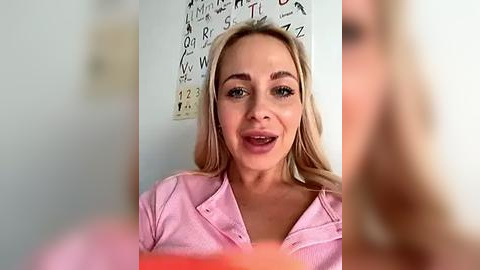 Media: Video of a smiling, blonde Caucasian woman with light skin, wearing a pink button-up shirt, standing in front of a wall adorned with a large alphabet chart.