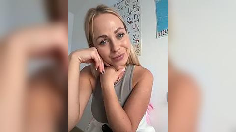 Media: A video of a smiling, fair-skinned blonde woman with long hair, wearing a sleeveless grey top, in a well-lit bedroom with a blue wall and a white bed.