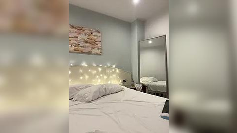 Media: Video of a minimalist bedroom with a tufted headboard, white bedding, a large mirror, and a wall-mounted abstract painting. The room has soft lighting, creating a serene ambiance.