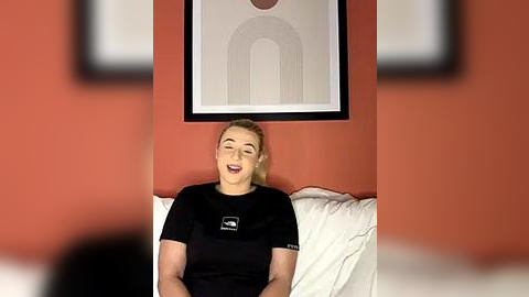 Media: Video of a smiling woman with blonde hair, wearing a black t-shirt, sitting on a white couch against an orange wall with abstract art.