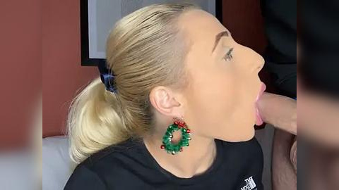 Media: Video of a blonde woman with a ponytail, wearing green and red earrings, performing oral sex on a man, in a modern room with white walls and framed art.