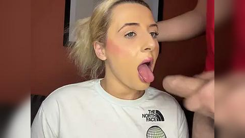 Media: Video of a blonde woman with fair skin, wearing a white t-shirt with \"The North Face\" logo, tongue out, in a red room.