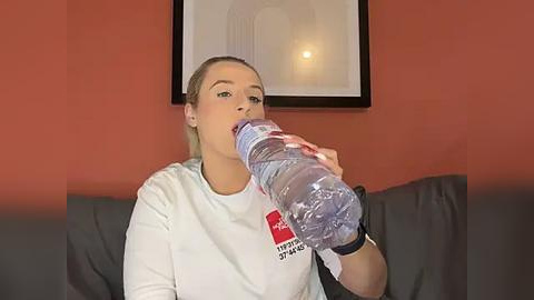 Media: Video of a blonde woman in a white shirt with \"Keep Calm and Carry On\" text, drinking from a large plastic water bottle on a gray couch against a red wall.