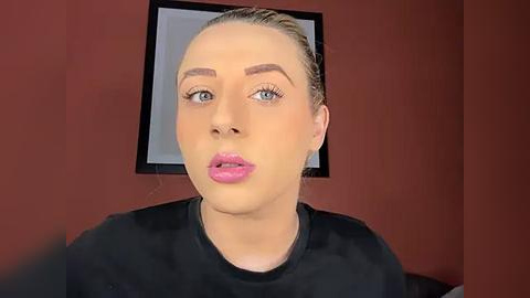 Media: Video of a woman with light skin and blonde hair, wearing black makeup, with pink lipstick and blue eye shadow, against a red wall with a framed mirror.