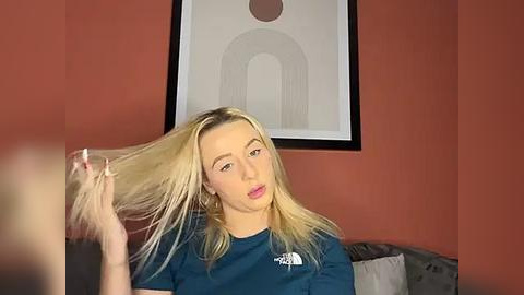 Media: Video of a blonde woman with fair skin and pink lipstick, holding her long hair. She wears a blue t-shirt. Background features a framed abstract art and a grey couch.