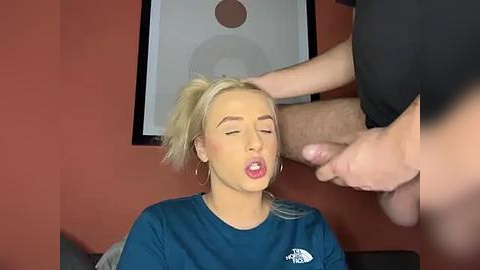 Media: A video depicts a blonde woman with fair skin, wearing a blue shirt, her mouth open in a suggestive expression. A man's erect penis is in the foreground. The background features a red wall with a circular abstract art piece.