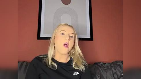 Media: Video of a blonde woman with fair skin, wearing a black sweatshirt, leaning forward on a grey couch. She has pink lips and is looking directly at the camera. Background includes a framed abstract art piece and reddish-orange wall.