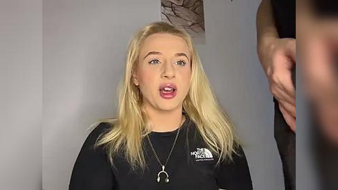 Media: A video of a blonde woman with fair skin, wearing a black top and necklace, mouth open in a surprised expression, indoors with blurred background.