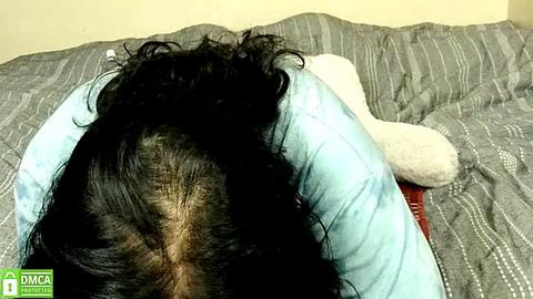 Media: Video of a person with long, dark hair lying face down on a bed with a gray striped blanket, wearing a light blue shirt.