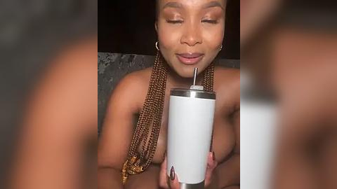 Media: Video of a topless, light-skinned Black woman with braided hair drinking from a white plastic cup. She's smiling, wearing hoop earrings, and has a soft, intimate lighting background.