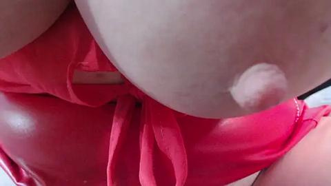 Media: A close-up video of a nude woman with fair skin, wearing a red satin bow tie. Her left breast is partially exposed, with a small, light-colored nipple visible. The background is blurred, focusing attention on the intimate detail.