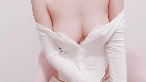 Media: Video of a fair-skinned woman wearing a white off-the-shoulder dress, exposing her ample cleavage. She has on white gloves and a subtle smile. The background is plain white, emphasizing the dress's texture.