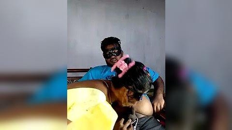 Media: Video of a young boy with dark skin and a pink headscarf, seated on a man's lap, wearing a blue shirt, in a dimly lit room with a concrete wall.