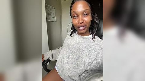 Media: Video of a young Black woman with short, black hair, wearing a gray sweater, sitting at a desk with a black chair. Background includes a whiteboard and a blurred person.