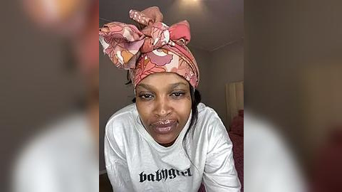 Media: Video of a pregnant woman with a brown complexion wearing a colorful, patterned headwrap, a white long-sleeve shirt with \"Baby\" printed on it, and a serious expression.