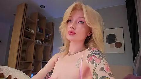 Media: Video of a blonde, pale-skinned woman with tattoos, wearing pink lingerie, standing indoors, surrounded by wooden shelves and abstract art.