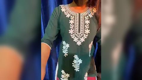 Media: Video of a person in a green kurta with intricate white embroidery, standing against a blue curtain. The person's face is partially obscured.